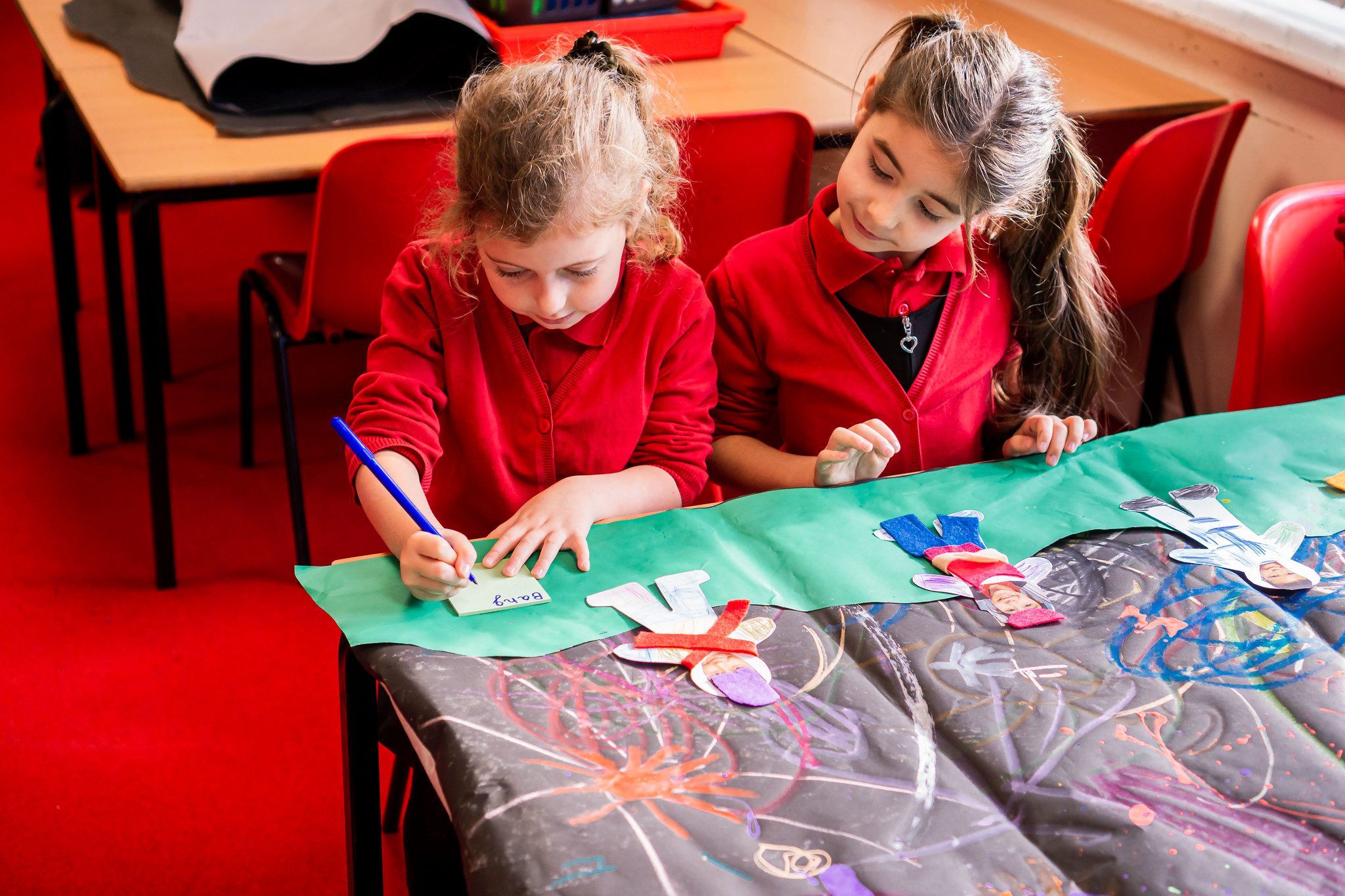 Two young students at desk, one with a pen in their writing the words 'Baby' on a post stick note hand and another seated and watching